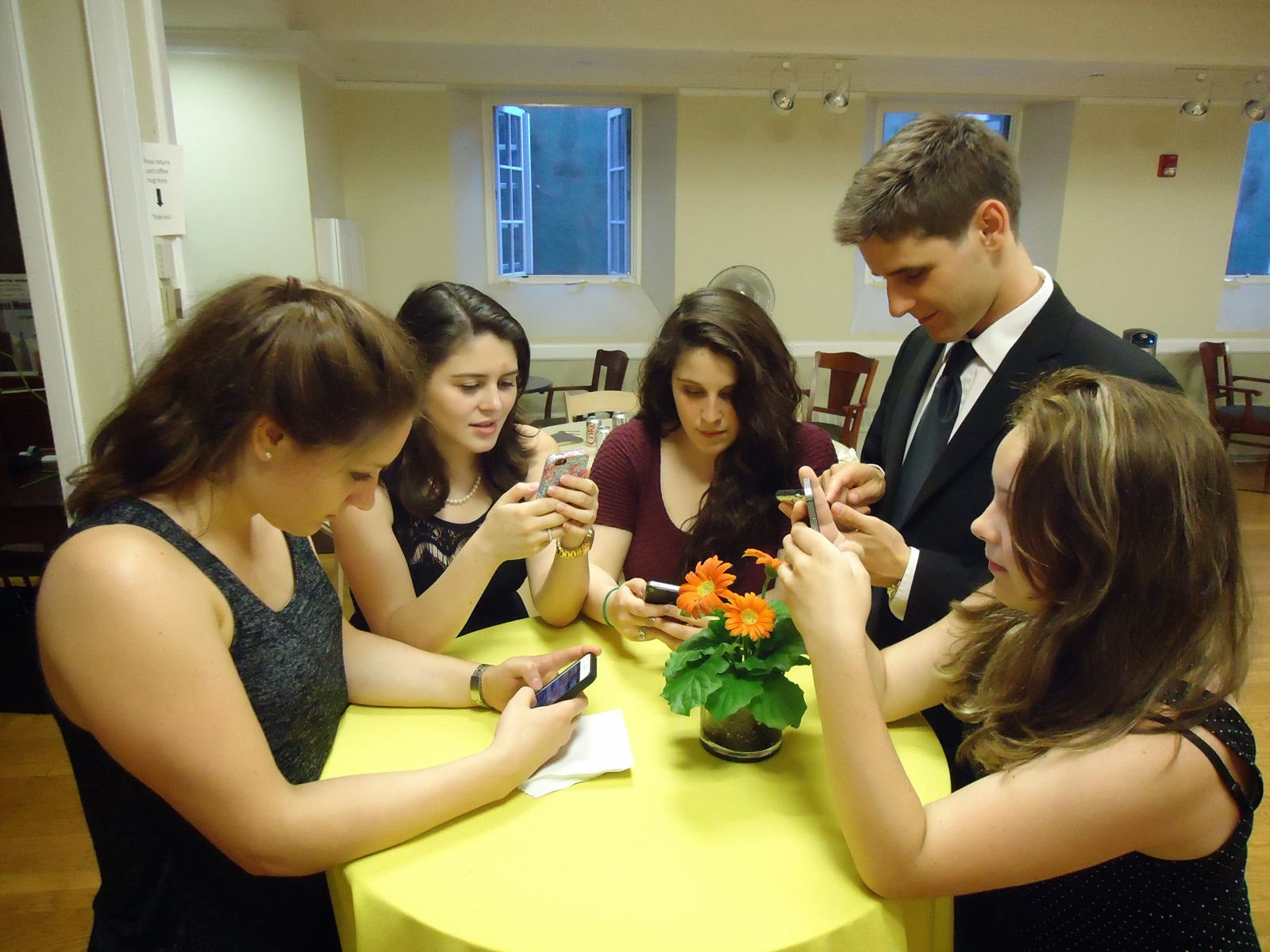 Five young people standing around a small yellow table, all looking down at their phones, with a pot of orange flowers in the middle.