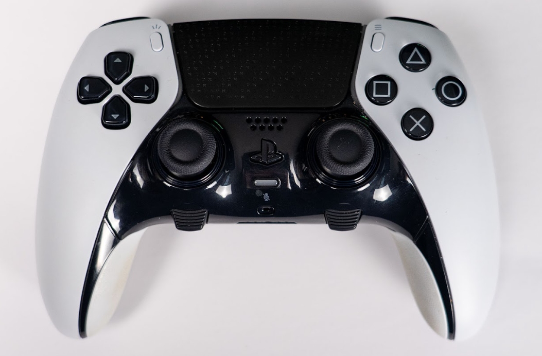 A white and black PlayStation DualSense Edge controller seen from above on a white surface, showing its touchpad, sticks and face buttons.