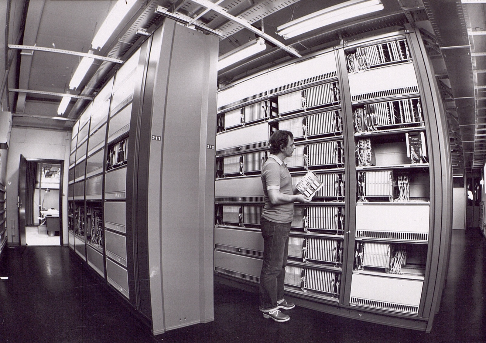 A black and white photo of a technician holding a circuit board between tall racks of telephone exchange equipment.