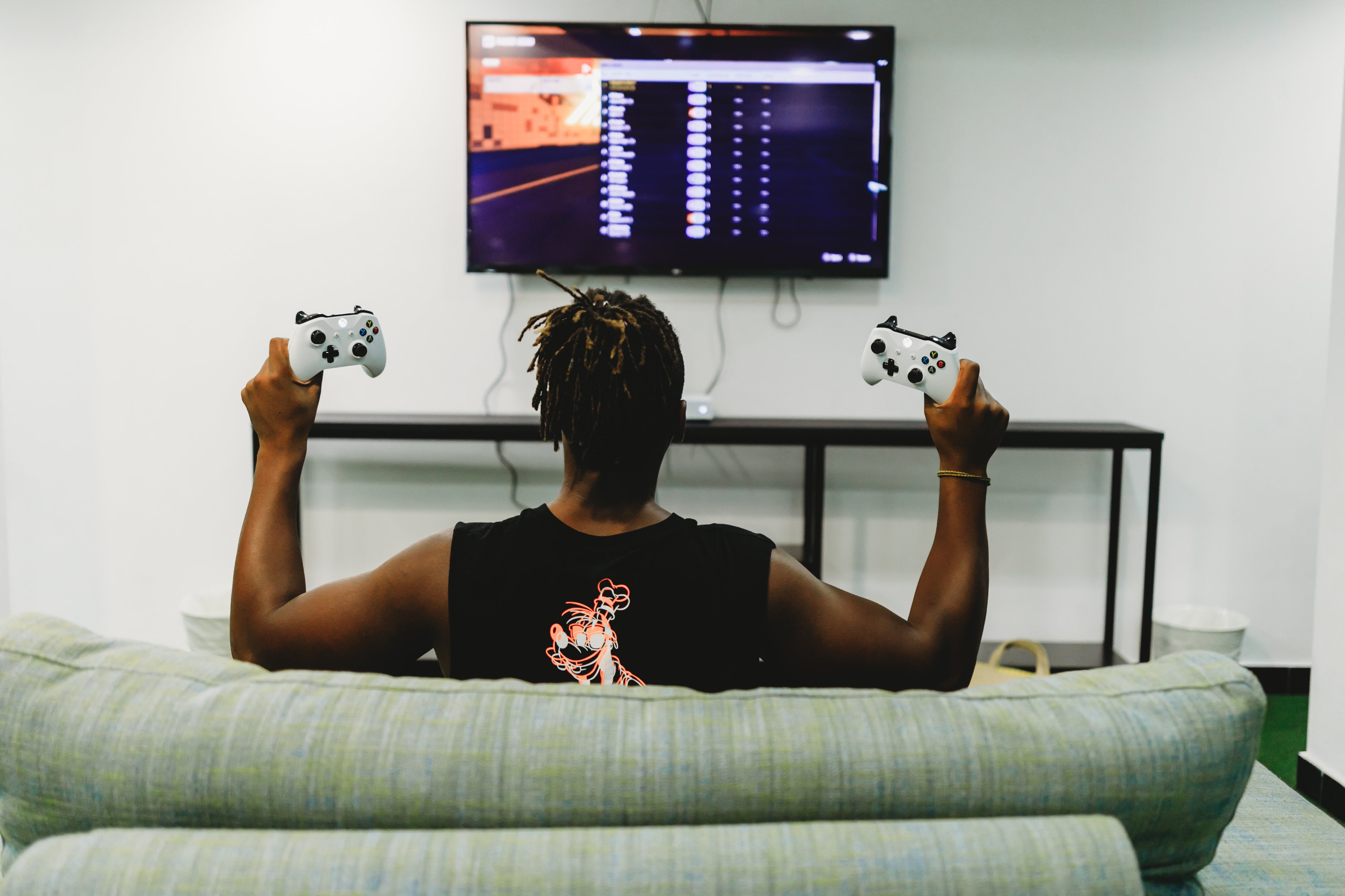 A player on a sofa, seen from behind, raises two white Xbox controllers toward a wall mounted TV showing a game menu.